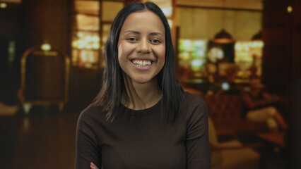 Hispanic woman with arms crossed smiles confidently indoors at a stylish hotel lounge, creating a...