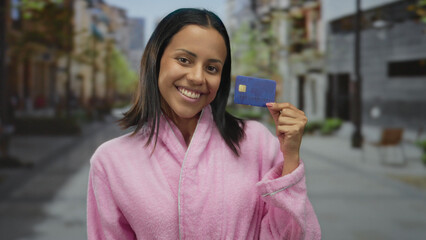 Young hispanic woman in a pink bathrobe holds a credit card smiling confidently in an outdoor urban...