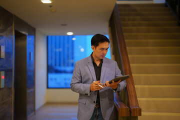 Young, focused Asian businessman using tablet and stylus pen, standing in a modern office lobby near the stairs, possibly reviewing documents or preparing for a meeting