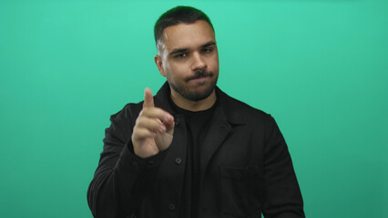 Young hispanic man wearing a black jacket with crossed arms against a vibrant green studio wall;...