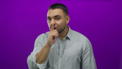 Young hispanic man smiling with finger to lips for silence in purple studio setting; mystery playfulness.