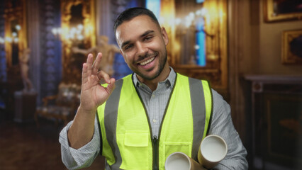 Young hispanic engineer holds rolled documents and makes ok sign in museum building; approval...