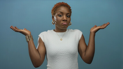 Woman shrugging shoulders and lifting both palms in a studio setting against a solid blue wall;...