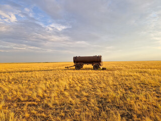 Fototapeta premium An old rusty water tank trailer stands alone in a vast golden steppe under a dramatic sky. Concept of solitude, abandonment, and rural landscape.