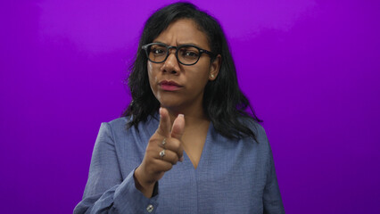 African american woman wearing glasses and a blue shirt frowns and points finger in studio; warning.