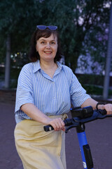 Smiling woman wearing a striped shirt and sunglasses rides an electric scooter along a park...