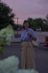 Woman standing outdoors near blurred foliage, smiling and enjoying the twilight atmosphere. She is...