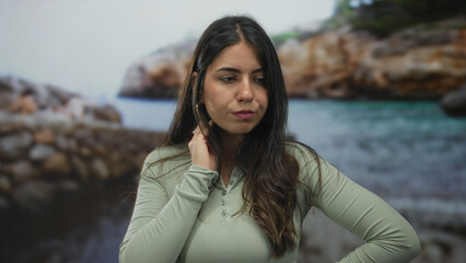 Young hispanic woman standing at seaside beach gazing pensively out towards the serene ocean under...