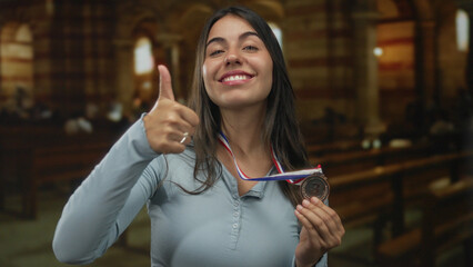 Woman pointing finger to bronze medal draped around her neck inside a church with wooden pews and...