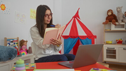 Hispanic woman teacher engaged in online teaching from a colorful kindergarten classroom filled...