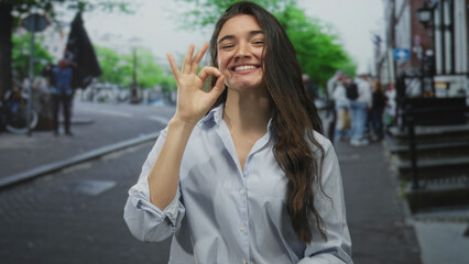 Young hispanic woman smiling and making ok sign with hand while standing on busy urban street lined...
