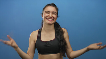 Fototapeta premium Woman wearing black sports bra raising arms with clenched fists and smiling face in blue studio; victory excitement.