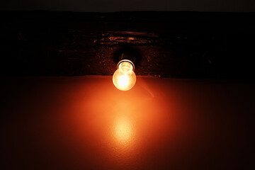 Close-up of glowing light bulb mounted to a wooden beam, casting a strong orange reflection on the...