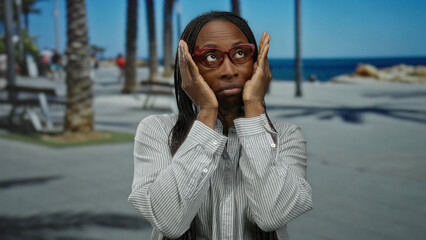Woman with curly hair wearing glasses makes playful expressions on a sunny seaside promenade,...