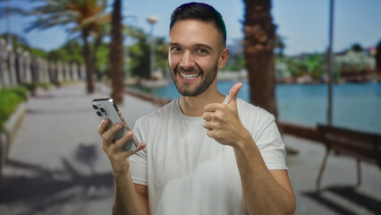 Hispanic man on promenade by seaside giving thumbs up while holding smartphone outdoors on sunny...
