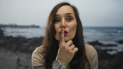 Young hispanic woman wearing beige sweater places finger to lips for silence at seaside; silence...