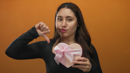 Young hispanic woman holds pink heart shaped gift box while pointing thumb down in studio;...