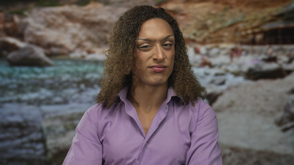 Transgender woman crosses arms at rocky beach wearing violet blouse under sunlight with curly hair;...