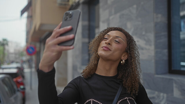Tall transgender woman holds smartphone at arm’s length smiling on street wearing hoop earrings; confidence. - Powered by Adobe