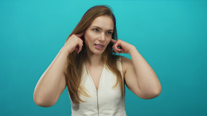 Fototapeta premium Young caucasian woman in white top covers ears with fingers in blue studio; calm concentration inner peace.