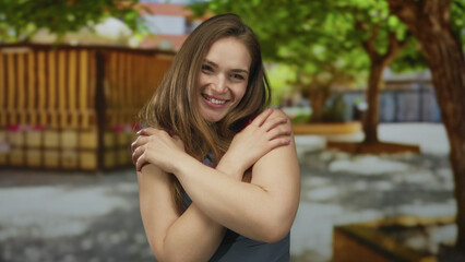 Woman smiling hugging herself with arms crossed in a green outdoor building setting; self love serenity.