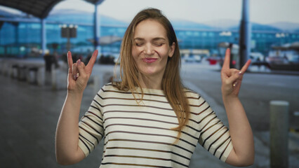 Fototapeta premium Young smiling caucasian woman in striped shirt raises both hands in rock horns gesture at airport terminal; excitement.