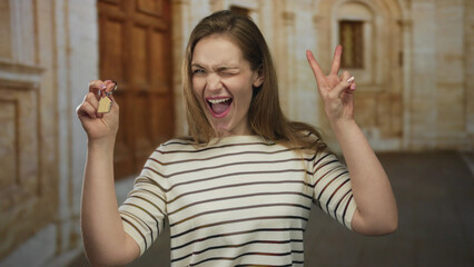 Woman holds house key and winks while making victory sign in front of building; home celebration...