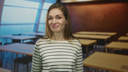 Young caucasian woman pointing finger at unoccupied wooden desk in bright classroom building;...