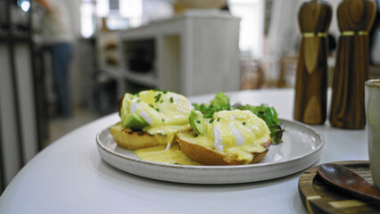 Eggs benedict on a table in a busy cafeteria featuring a blurred person in the background and pepper mills on the table.