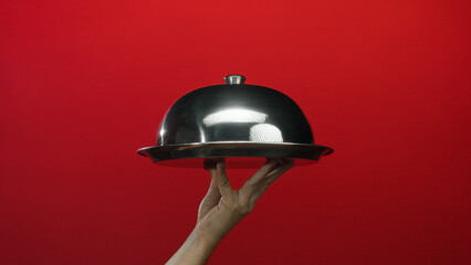 Hand holding a shiny cloche against a vibrant red background, showcasing a metallic serving...