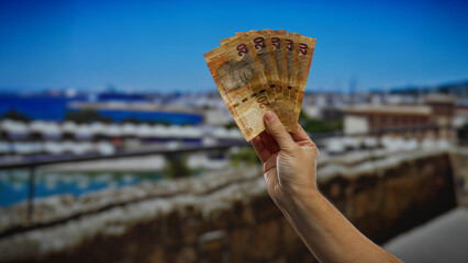 Hand holding south african rand banknotes with a city seaside port view, illustrating summer...