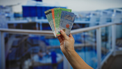 Hand holding malaysian ringgit in seaside port, showcasing colorful banknotes on a boat background,...