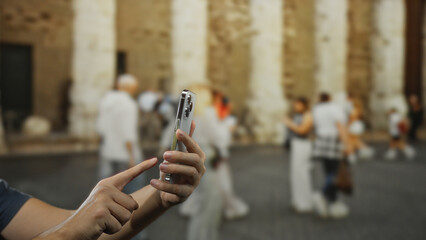Man using smartphone outdoors on city street capturing moment, with focus on hand and device, people blurred in background for urban lifestyle context.