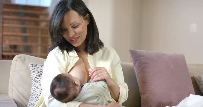 Mid adult woman unbuttoning yellow shirt, feeding infant in onesie on sofa with mauve pillow