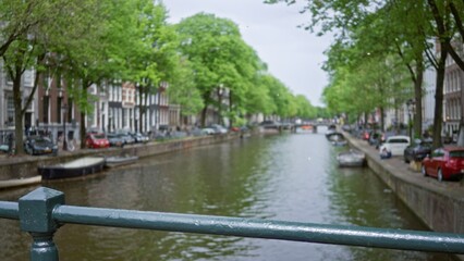 Defocused view of amsterdam canals featuring blurred canal, bridge railings, and vibrant green trees along the historic dutch cityscape under soft daylight.