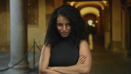 Young hispanic woman sticks tongue out in old town street archway while crossing arms; playful...