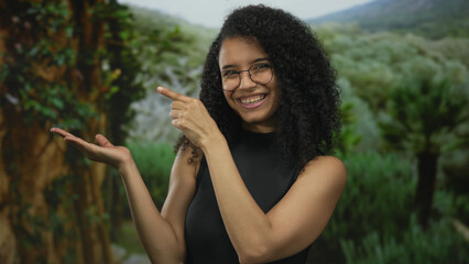 Woman wearing glasses and black sleeveless top smiles and points finger to palm in green forest;...