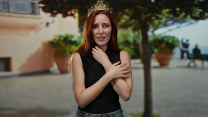 Woman with red hair and crown looks anxious in city street setting with trees.