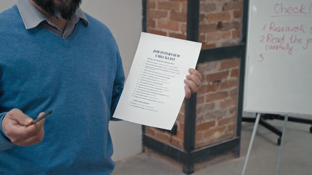 Medium shot of middle-aged Caucasian male teacher in blue sweater holding job interview checklist while training inmates in basic social skills during rehabilitation course in prison