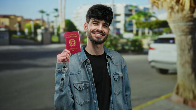 Fototapeta Confident man holding italian passport smiles on urban street with palm trees and buildings under blue sky.