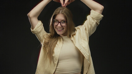 Blonde woman wearing glasses and smiling while forming heart shape with bare hands in studio under...