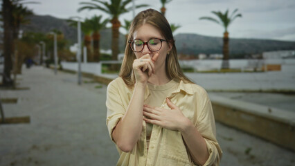 Woman with glasses and beige shirt holds hand on chest walking along palm lined street by...