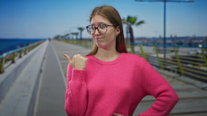 Woman finger pointing to object on street promenade by seaside in pink sweater with skeptical...