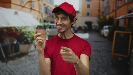 Smiling young delivery man holding disposable coffee cup points finger to cup on urban street;...