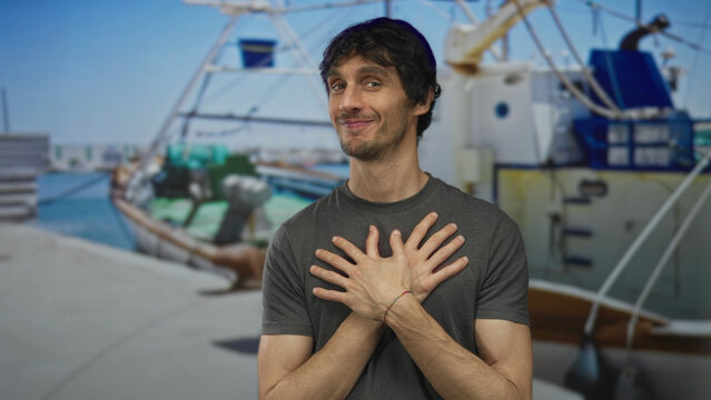 Man with hand on chest next to fishing boat on street dock under clear blue sky wearing grey t shirt and smiling gently; gratitude sincerity.