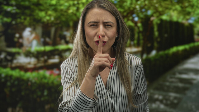 Blonde woman finger to lips for silence beneath sunlit leafy canopy beside brick pathway in park; secrecy privacy intrigue.