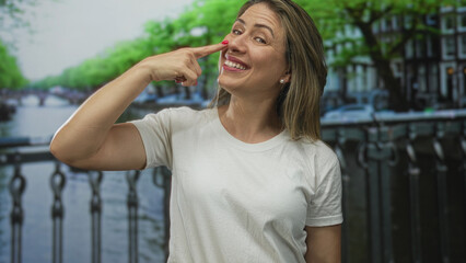 Woman in white t shirt points finger to nose beside canal railing on a city street lined with leafy trees; playfulness.