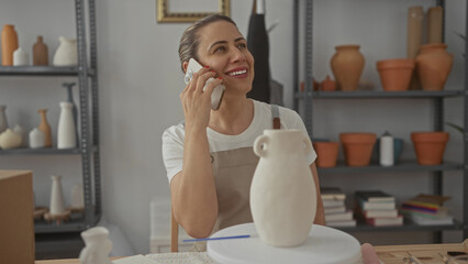Woman smiling holds smartphone to ear beside ceramic vase on pottery wheel on work table in studio; contentment.
