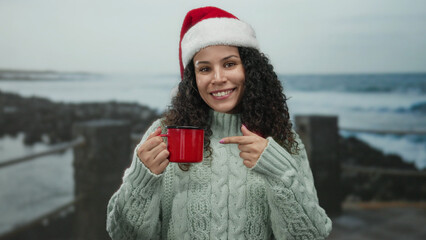 Obraz premium Woman wearing santa hat points at red mug by the beach, showing joy by the sea in a casual sweater, blending holiday cheer with coastal relaxation.