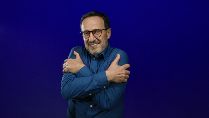 Mature hispanic man smiling and hugging himself in a blue shirt against an isolated blue background, embodying happiness and warmth.